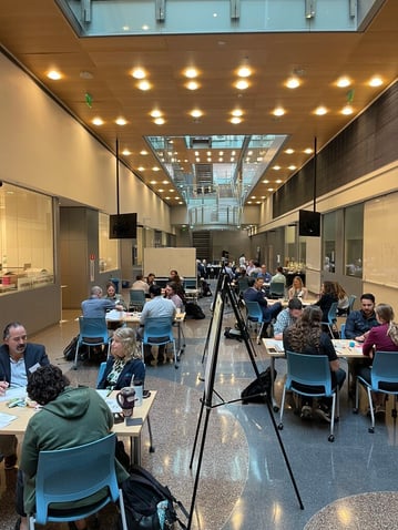 An image of the workshop hall, where small groups are gathered around tables and whiteboards discussing their work.