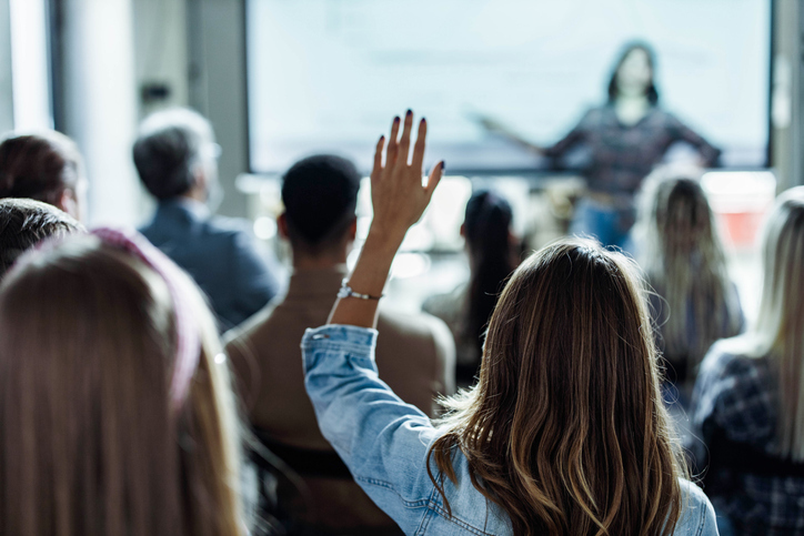 Back view of a white woman raising her hand at a presentation.