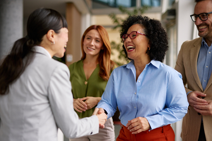 A Black woman and an Asian woman shake hands with other colleagues around.