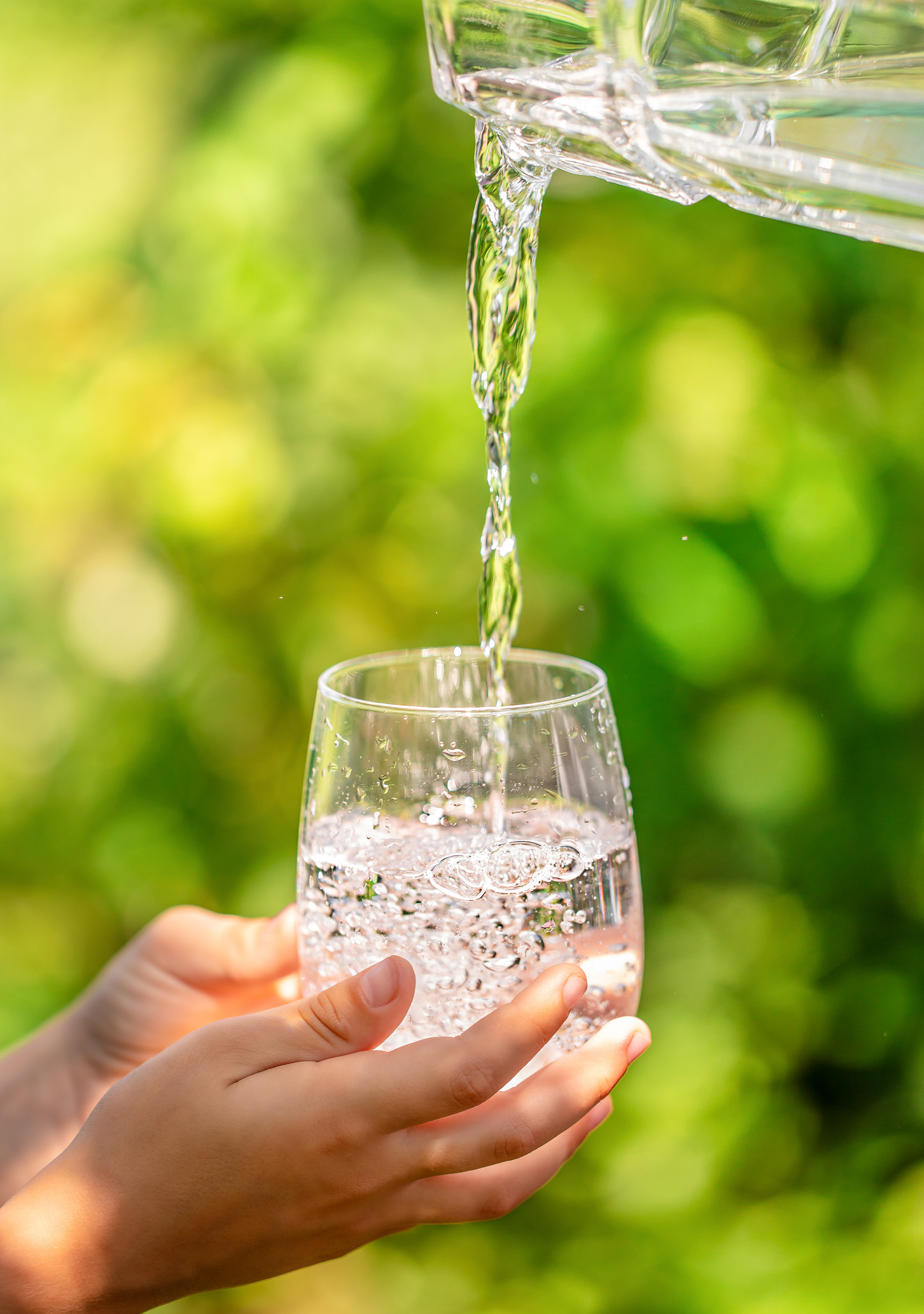 Fresh drinking water pouring into glass outdoors with human hands holding the cup.